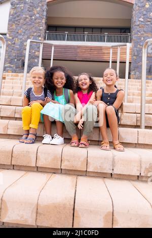 Die ganze Länge der multirassischen fröhlichen Grundschüler sitzen auf Stufen gegen das Schulgebäude Stockfoto