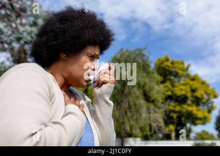 afroamerikanische Frau mit mittlerem Erwachsenen und Hand auf der Brust, die Asthma-Inhalator verwendet, während sie im Freien steht Stockfoto