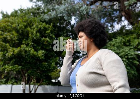 Seitenansicht einer afroamerikanischen Frau mit mittlerem Erwachsenen, die Asthma-Inhalator verwendet, während sie gegen Bäume steht Stockfoto