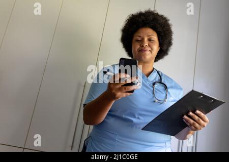 afroamerikanische Krankenschwester mit mittlerem Erwachsenen, die Smartphone benutzt, während sie die Zwischenablage im Krankenhaus hält Stockfoto