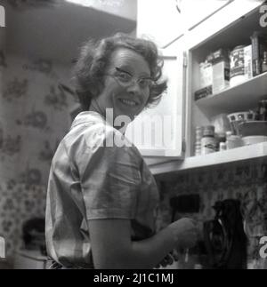 1959, historisch, eine Dame in ihrer Küche, in einem offenen Schrank, Stockport, Manchester, England, Großbritannien. Sie trägt eine geflügelte Brille, die in dieser Zeit modisch ist. Stockfoto