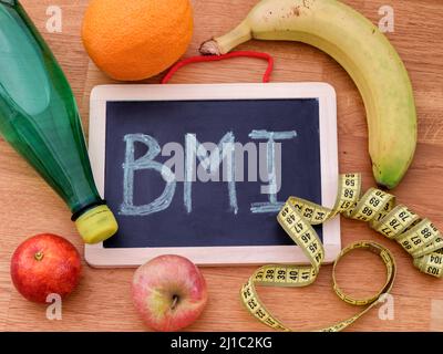 Die Abkürzung BMI (Body Mass Index) auf einer Tafel und einigen Früchten, einer Wasserflasche und einem Maßband um sie herum. Nahaufnahme. Stockfoto