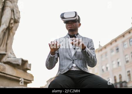 afroamerikanischer Mann im stilvollen Business-Anzug, der auf der Stadtstraße sitzt und VR-Brille verwendet. Konzept von Menschen, Innovation und modernen Lebensstilen. Stockfoto