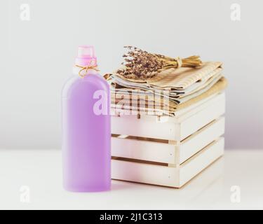 Kunststoffverpackung mit natürlichem Flüssigwaschpulver oder Leinenaufbereiter mit Küchentüchern und Lavendelblüten auf einem weißen Tisch. Nachbau von Organi Stockfoto