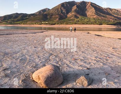 Zwei weit entfernte Menschen stehen auf dem Grund eines ausgetrockneten Flussbettes vor dem Hintergrund eines Sees und von Bergen. Konzept der kleinen Menschen, große Welt. Stockfoto