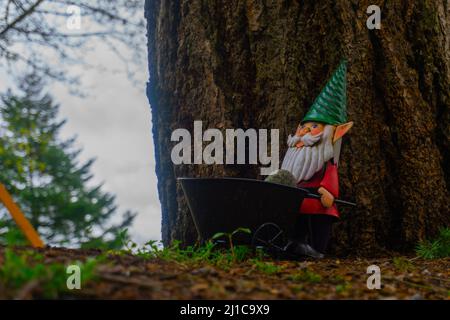 Ein Gartenzwerg hält eine Schubkarre vor einem Baum mit einem weiteren Baum im Hintergrund. Stockfoto