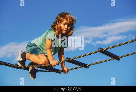 Kind klettert im Netz. Netter Junge klettert die Leiter auf dem Spielplatz hoch. Kind klettert die Leiter gegen den blauen Himmel hinauf. Stockfoto