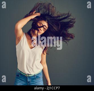 Ihr Haar hat einen eigenen Geist. Studioaufnahme einer unbeschwerten jungen Frau, die ihre Haare hält, während es vom Wind geblasen wird, während sie gegen eine Dunkelheit steht Stockfoto