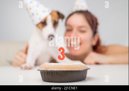 Kaukasische Frau und Jack russell Terrier in Urlaubsmützen betrachten den Kuchen mit einer Kerze. Der Hund und der Besitzer feiern den dritten Geburtstag. Stockfoto