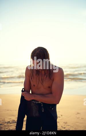 Machen Sie sich bereit, in den blauen Himmel zu gehen. Aufnahme eines jungen Mannes, der am Strand einen Neoprenanzug anlegt. Stockfoto