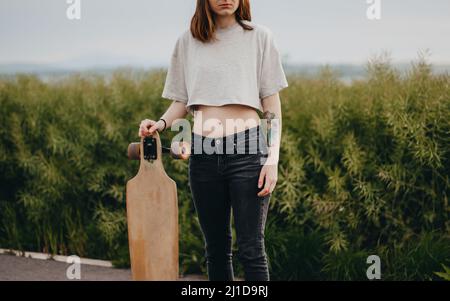 Vertikales Plakat. Junge gesichtslose Frau hält ein Longboard in der Hand. Im Hintergrund ist grüne Natur Stockfoto