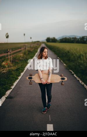 Vertikales Poster. Das Mädchen hält ein Longboard in der Hand auf einer unbefestigten Straße und schaut weg Stockfoto