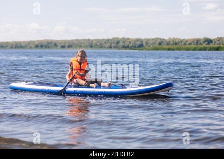 Kleine blonde Mädchen Pfütze Boarding auf schönen See Rudern mit Ruder in den Händen Blick auf rippled Wasser in Weste Schwimmweste. Aktivurlaub. Einimulation Stockfoto