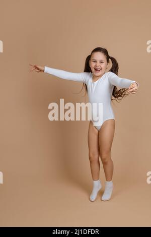 Porträt von jungen niedlichen glücklich lächelndes Mädchen mit langen dunklen Haaren in weißen Trainingsanzug und Socken springen auf braunem Hintergrund, die Hände zu verbreiten Stockfoto