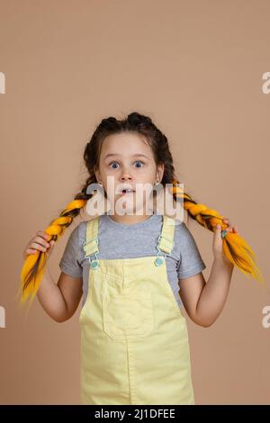 Urkomische kleine Hündin mit gelben Kanekalon-Zöpfen, die mit verblüfften, überraschten Augen mit geöffnetem Mund und gelbem Gesicht auf die Kamera blicken Stockfoto