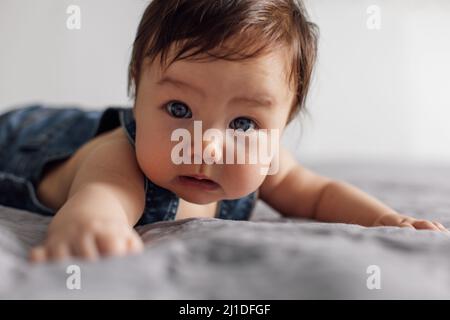 Portrait eines hübschen Babys, das Denim-Strampler trägt und zu Hause auf einer grauen Decke liegt. Erschrocken blauäugig Kleinkind kriechen auf dem Bett im Schlafzimmer, erkunden Welt Stockfoto