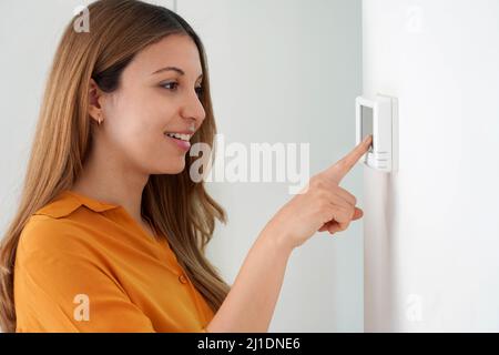 Porträt einer schönen Frau, die die Temperatur senkt, um Energie zu sparen. Frau, die zu Hause den digitalen Thermostat für die Zentralheizung justiert. Stockfoto