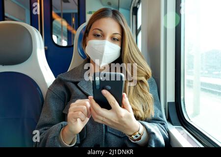 Entspannte Frau mit KN95 FFP2 Gesichtsmaske mit Smartphone-App. Zug Passagier mit Schutzmaske Reisen sitzen SMS auf Handy. Reise s Stockfoto