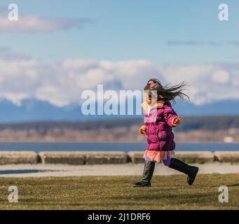 Kleines niedliches Mädchen in einem rosa Mantel, das auf dem Gras läuft. Glückliches kleines Mädchen in Herbstlandschaft. Selektiver Fokus, Kopierbereich für Text-März 15,2021-Vancouvere Stockfoto