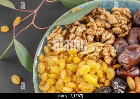 Goldene Rosinen, Walnüsse, Mandeln, Pflaumen und Datteln in der blauen Schale auf schwarzem Hintergrund mit einem Zweig mit grünen Blättern. Arabisches Gericht mit Nüssen und dr Stockfoto