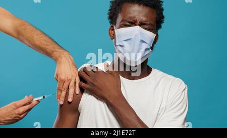 Verängstigte Person mit Gesichtsmaske, die gegen Coronavirus geimpft wird. Hände des Arztes mit Spritze, die jungen Mann mit Schutzmaske impfen. Erwachsene Angst vor Nadel und Impfung. Stockfoto