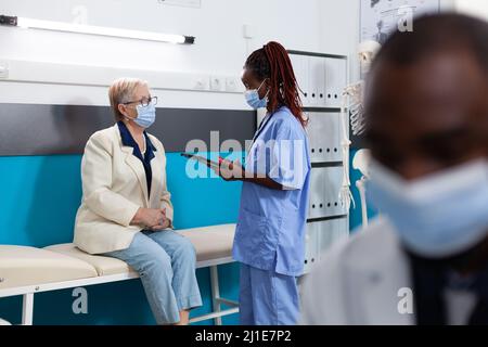 afroamerikanische Krankenschwester, die eine schützende Gesichtsmaske trägt, um eine Infektion mit dem Coronavirus zu verhindern und die Krankheitsdiagnose mit einer pensionierten Frau zu besprechen. Arzthelferin, die die medikamentöse Behandlung auf eine Tablette schreibt Stockfoto