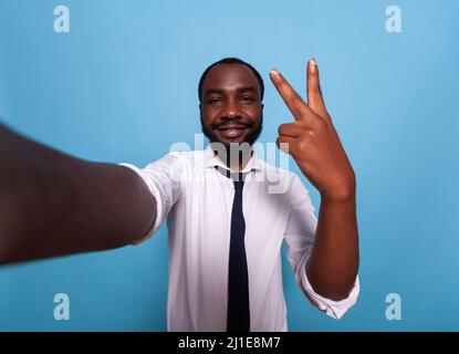 Lächelnder Vlogger zeigt auf blauem Hintergrund ein Friedenszeichen an der Kamera in einer Videokonferenz. Weitwinkel pov von glücklichen Influencer, die ein Smartphone Selfie mit zwei Fingern Geste für Social Media heben. Stockfoto