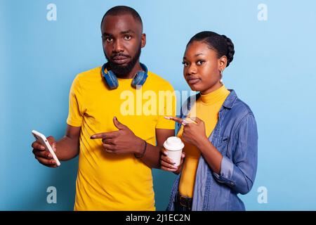 afroamerikanische Liebhaber zeigen auf Smartphone-Bildschirm im Studio, Werbetechnik und modernes Handy. Mann und Frau, die mit dem Zeigefinger auf das Mobiltelefon zeigen und lächeln. Stockfoto