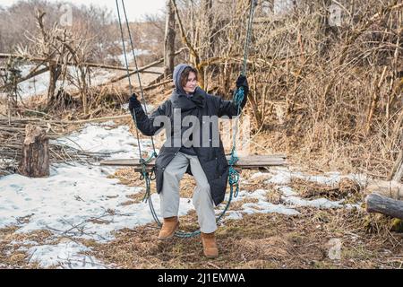 Teenager Mädchen im Winter warme Kleidung reitet auf Schaukel auf langen Seil an Baum in ländlicher Umgebung gebunden. Echtes Landleben Stockfoto