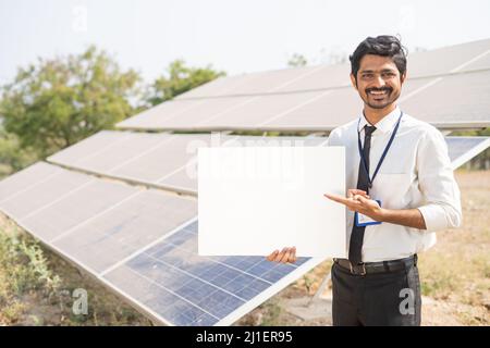 Lächelnder Bankbeamter zeigt weißes leeres Brett, indem er vor dem Solarpanel auf Ackerland die Kamera anschaut - Konzept von Botschaft, Werbung, Finanzen Stockfoto