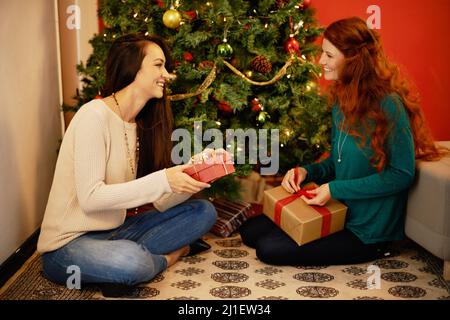 Können Sie die Weihnachtsfreude spüren. Aufnahme von zwei jungen Frauen, die an einem Weihnachtsbaum sitzen und ihre Geschenke öffnen. Stockfoto