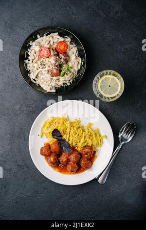 Zwei Teller Pasta mit Fleischbällchen aus Fleisch auf pflanzlicher Basis, Pilzen, Basilikum, Tomaten- und Rahmsoße, Tomaten auf dunklem Betonboden. Stockfoto
