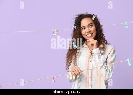 Nachdenkliche junge afroamerikanische Frau mit hängenden Wäscheklammern auf lila Hintergrund Stockfoto