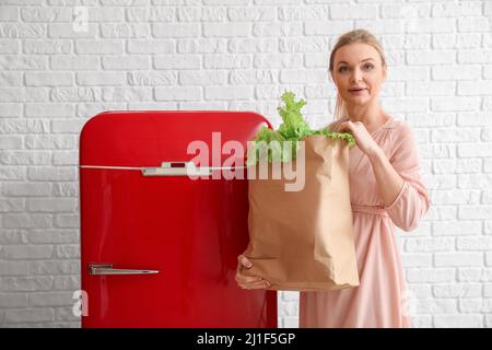 Frau mit Beutel mit frischen Produkten in der Nähe von stilvollen roten Kühlschrank auf weißem Backstein Wand Hintergrund Stockfoto
