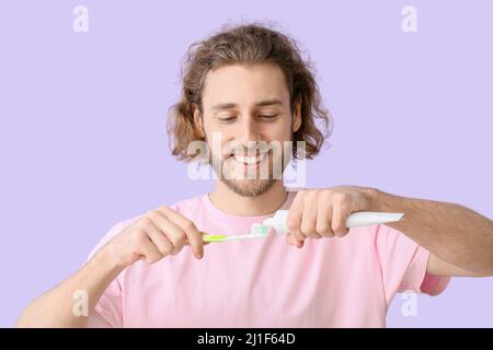 Junger Mann, der Zahnpasta auf den Pinsel auf fliederem Hintergrund aufsetzt Stockfoto