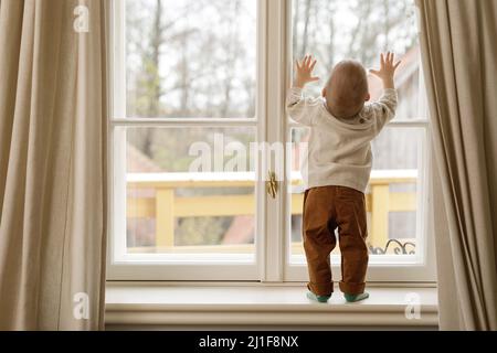 Der kleine Junge steht auf der Fensterbank und schaut auf das Fenster Stockfoto