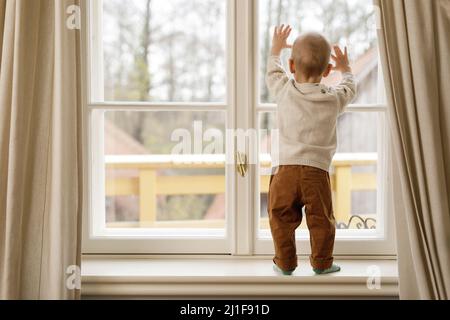 Der kleine Junge steht auf der Fensterbank und schaut auf das Fenster Stockfoto