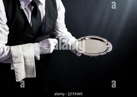 Porträt eines Butlers oder Kellers in schwarzer Weste und weißen Handschuhen mit Serviertablett auf schwarzem Hintergrund. Dienstleistungsbranche und professionelle Höflichkeit. Stockfoto