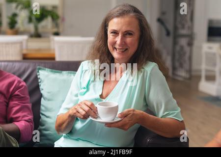 Porträt einer lächelnden kaukasischen älteren Frau, die Kaffee getrunken hat, während sie von einem Freund zu Hause sitzt Stockfoto