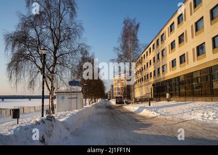 Rovaniemi, Finnland - 17.. März 2022: Eine verschneite Straße und Gebäude erstrecken sich am Ufer des gefrorenen Kemijoki-Flusses, Rovaniemi, Finnland. Stockfoto