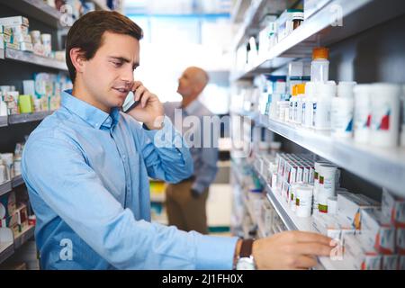 Wie lautet der Name wieder? Ausgeschnittene Aufnahme eines hübschen jungen Mannes, der einen Telefonanruf abgibt, während er in einer Apotheke nach Medikamenten sucht. Stockfoto