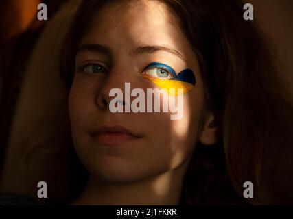 Nachdenkliche Gesicht einer jungen Frau aus der Ukraine mit gelb-blauem Herz um das Auge, hervorgehoben durch Lichtstreifen aus Schatten. Verlust der Hoffnung und Sadne Stockfoto