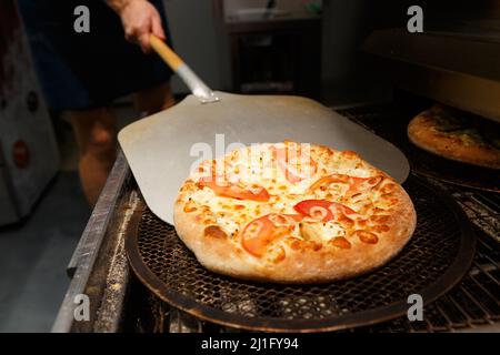 Ein Arbeiter nimmt in einem Restaurant frisch gebackene Pizza aus dem modernen Transportofen. Stockfoto