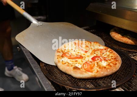 Ein Arbeiter nimmt in einem Restaurant frisch gebackene Pizza aus dem modernen Transportofen. Stockfoto