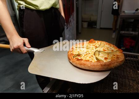 Ein Arbeiter nimmt in einem Restaurant frisch gebackene Pizza mit Gurken und Mozzarella aus dem modernen Transportofen. Stockfoto