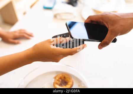 Verkürzte Hände von multirassischen Kunden und Besitzer tun kontaktlose Zahlung über Smartphone im Café. Unverändert, Café-Kultur, Technologie, Beruf Stockfoto