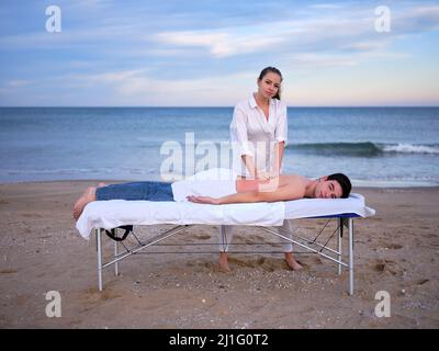 Ein junger Therapeut, der eine Chiromassage an einem jungen Mann zur Behandlung einer Lendenwirbelverletzung im Freien durchführt, und dabei auf einen Strand in Valencia blickt. Stockfoto