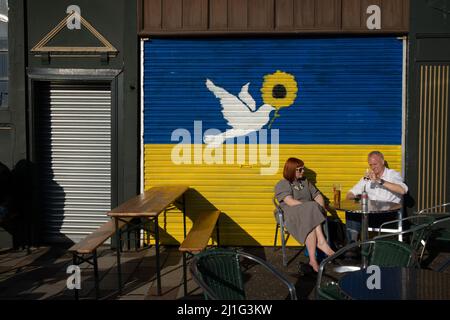 Glasgow, Großbritannien, 25.. März 2022. Ein Paar genießt die Sonne vor einem Wandgemälde an der Wand des Teesaals von BiddyÕs, das die ukrainische Flagge und eine Friedenstaube zeigt, gemalt, um die Unterstützung der Ukraine in ihrem andauernden Krieg gegen die Invasion Russlands am östlichen Ende von Glasgow, Schottland, zu zeigen. 25. März 2022. Foto: Jeremy Sutton-Hibbert/Alamy Live News. Stockfoto