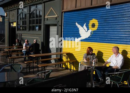 Glasgow, Großbritannien, 25.. März 2022. Ein Paar genießt die Sonne vor einem Wandgemälde an der Wand des Teesaals von BiddyÕs, das die ukrainische Flagge und eine Friedenstaube zeigt, gemalt, um die Unterstützung der Ukraine in ihrem andauernden Krieg gegen die Invasion Russlands am östlichen Ende von Glasgow, Schottland, zu zeigen. 25. März 2022. Foto: Jeremy Sutton-Hibbert/Alamy Live News. Stockfoto