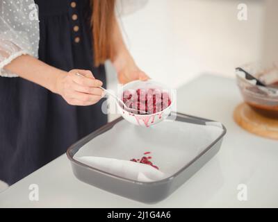 Bäckerei, Lebensmittel und Menschen Konzept. Frau bereitet Schokolade Brownie am Küchentisch, Nahaufnahme. Weibliche Köchin setzt Kirschen in Zucker auf ein Backblech Stockfoto